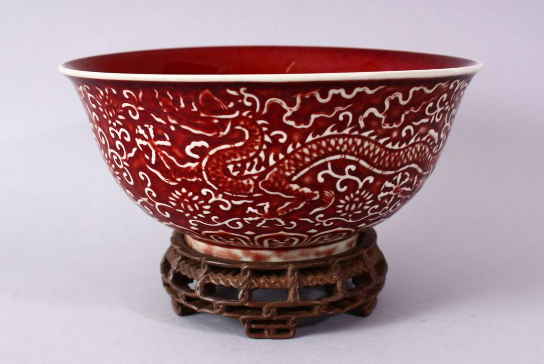 A CHINESE COPPER RED CARVED COPPER RED PORCELAIN DRAGON: A CHINESE COPPER RED CARVED COPPER RED PORCELAIN DRAGON BOWL, with decoration of dragins amongst flora, with a carved wooden stand, the underside with a six character mark, 21cm.