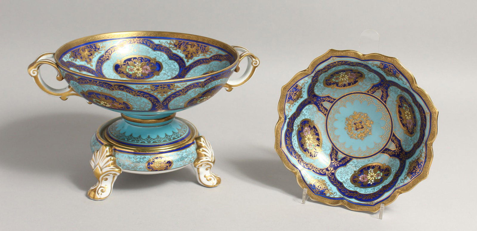 A NORITAKE PORCELAIN TWIN HANDLED PEDESTAL BOWL, pale: A NORITAKE PORCELAIN TWIN HANDLED PEDESTAL BOWL, pale blue ground with rich blue floral decorated panels, gilded borders, together with a matching stand and lobed bowl (2). 12ins and 8ins diameter.