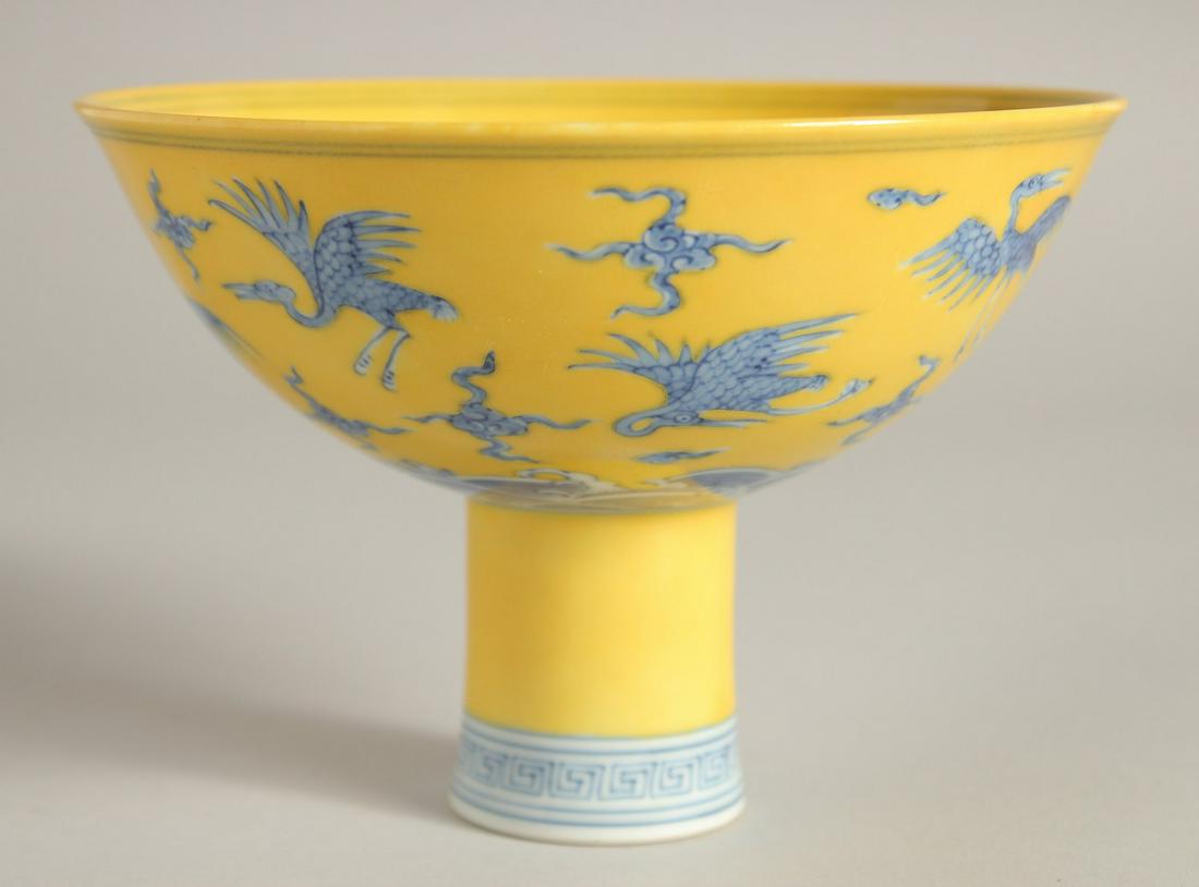A CHINESE YELLOW GROUND BLUE AND WHITE STEM CUP, the exterior decorated with cranes and stylised: A CHINESE YELLOW GROUND BLUE AND WHITE STEM CUP, the exterior decorated with cranes and stylised clouds and waves, the inner foot rim with six-character mark, bowl 15cm diameter.