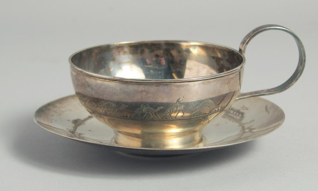 A LARGE IRAQI SILVER CUP AND SAUCER, with engraved decoration of camels and boats, weight 220g, (2: A LARGE IRAQI SILVER CUP AND SAUCER, with engraved decoration of camels and boats, weight 220g, (2 pieces).