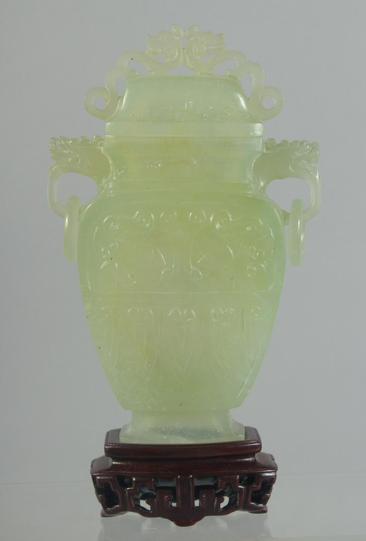 A CHINESE CARVED JADE LIDDED VASE AND WOODEN STAND, the vase with archaic style carving and drop: A CHINESE CARVED JADE LIDDED VASE AND WOODEN STAND, the vase with archaic style carving and drop ring handles, 18.5cm high (on stand).