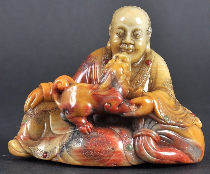 A FINE CHINESE SOAPSTONE CARVED FIGURE OF A SEATE: A FINE CHINESE SOAPSTONE CARVED FIGURE OF A SEATED SCHOLAR probably 18th century, well carved with a kylin upon his lap, with jewelled decoration. Natural fissures. 2. 75ins wide.