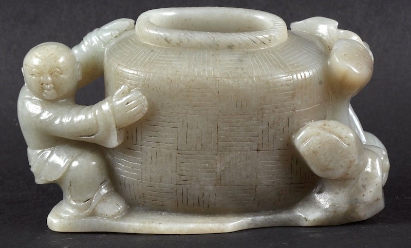 A CHINESE CARVED GREEN JADE INK POT formed as a young: A CHINESE CARVED GREEN JADE INK POT formed as a young boy climbing upon a basket, with foliate mounts. Fissures. 4.25ins wide.
