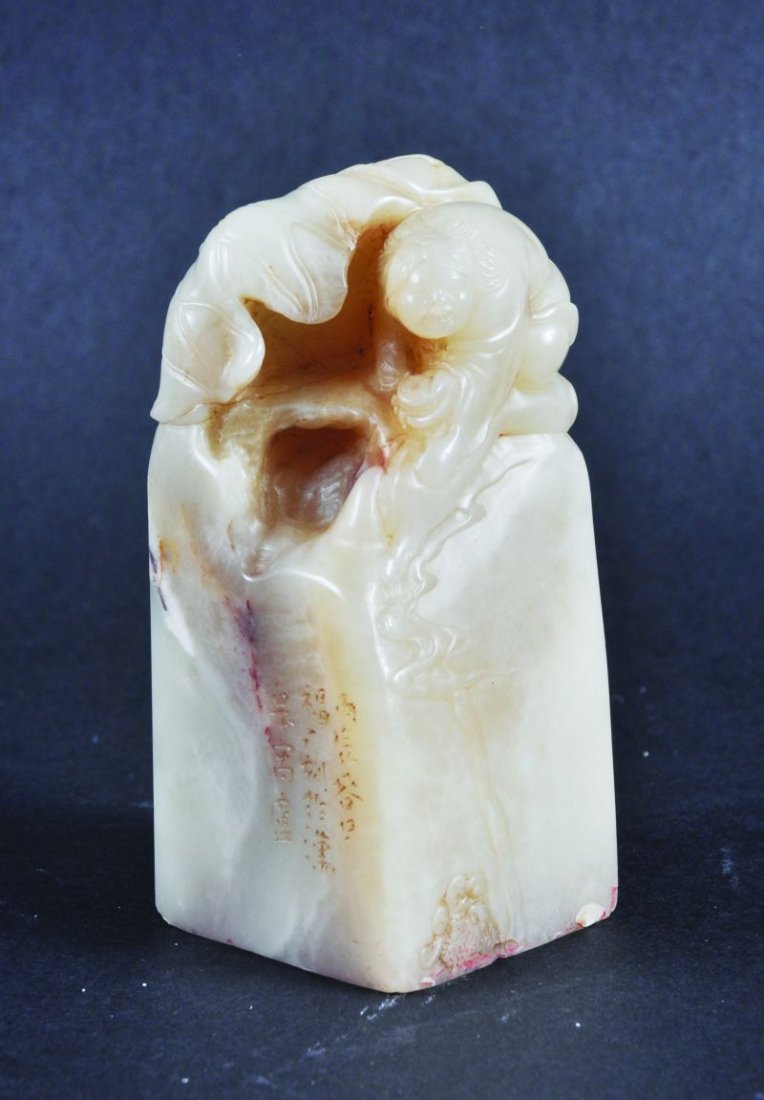 A CHINESE CARVED SOAPSTONE SEAL, finely decorated with: A CHINESE CARVED SOAPSTONE SEAL, finely decorated with a boy seated next to a canopy of lotus leaves, together with a fitted box, the seal 3.75in high.