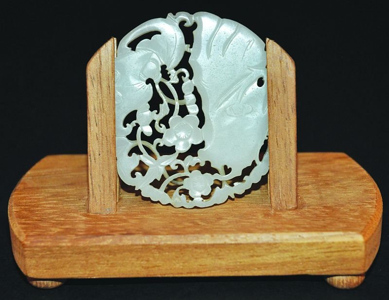 A SMALL 19TH/20TH CENTURY CHINESE JADE PLAQUE, with: A SMALL 19TH/20TH CENTURY CHINESE JADE PLAQUE, with wood stand, the carved and pierced decoration in the form of a bat in flight above foliage and rockwork, 2.25in wide & 1.8in high. (2) £200-300