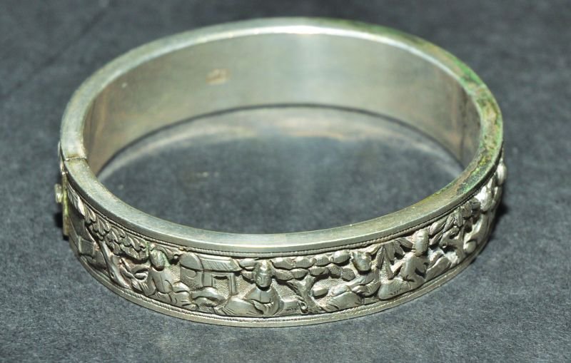 A CHINESE HINGED SILVER-METAL BANGLE BY WANG HING, of: A CHINESE HINGED SILVER-METAL BANGLE BY WANG HING, of oval form, the outer surface decorated with diminutive figures in a rock strewn garden setting, the inside rim with impressed ‘WH’, 2.5in x 2.
