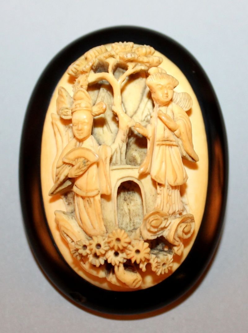 TWO 19TH CENTURY CHINESE CANTON IVORY OVAL BROOCHES,: TWO 19TH CENTURY CHINESE CANTON IVORY OVAL BROOCHES, one of them mounted on ebony and unusually carved in deep relief with a mandarin and lady in a garden setting, the other with a silver-metal mount,