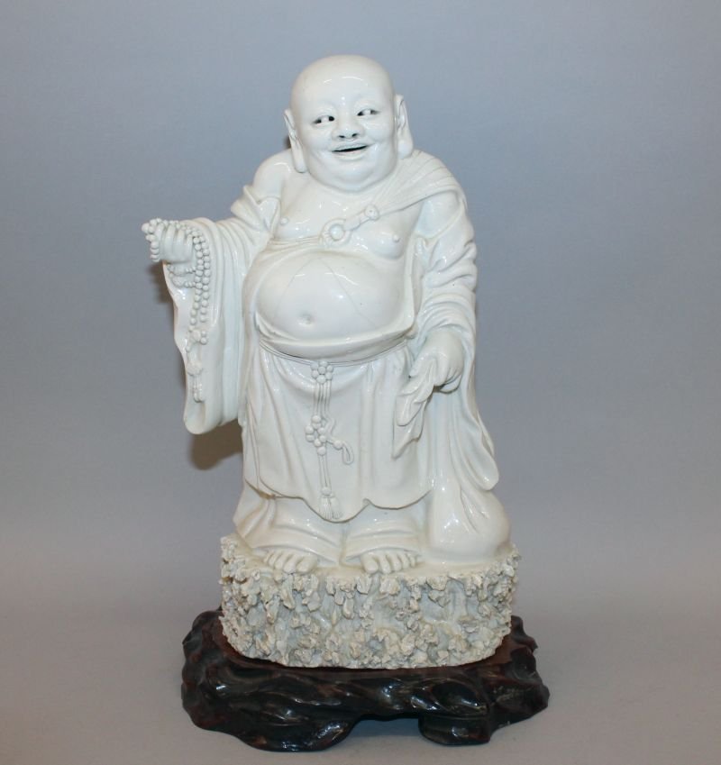 A 19TH CENTURY CHINESE BLANC-DE-CHINE PORCELAIN FIGURE: A 19TH CENTURY CHINESE BLANC-DE-CHINE PORCELAIN FIGURE OF BUDAI, together with a fitted wood stand, the deity standing on a rockwork base and holding a string of beads in one hand, 13.4in high overall