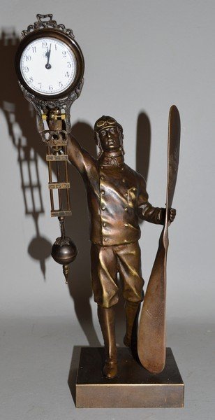 A BRONZED FLYING PILOT PENDULUM CLOCK holding a: A BRONZED FLYING PILOT PENDULUM CLOCK holding a propeller on square base. 11.5ins high.