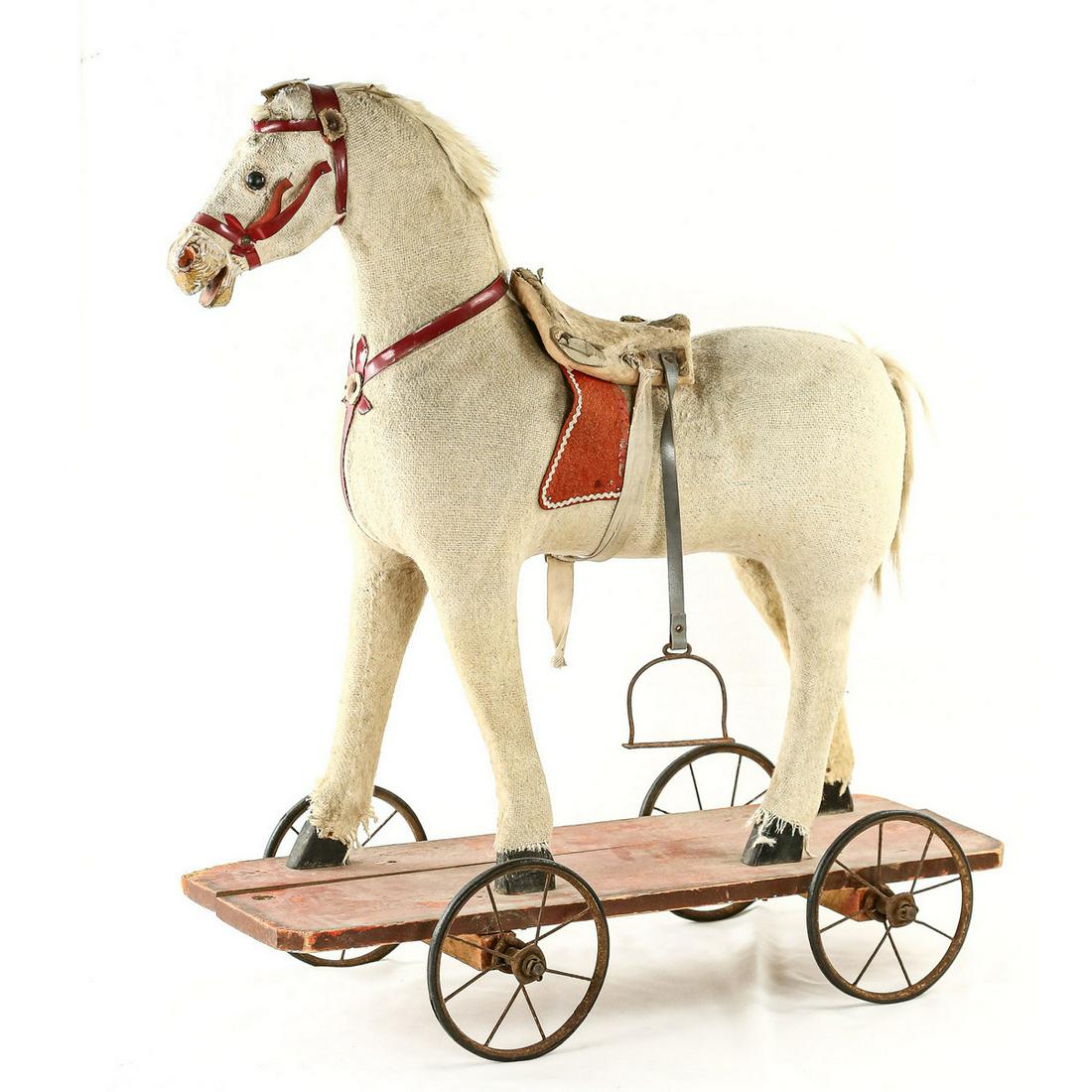 Hobby Horse: Circa 1900. Child's toy horse. Woven cloth body horse with button eyes, leather harness, felt covered saddle, missing one stirrup, animal hair mane and tail. Mounted on a wood plank with wire spoke wh