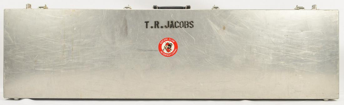 Hard Metal Gun Case: A hard metal gun case with four latches. Case is named to "T.R. Jacobs" and has a Safari Club International sticker on the side. Foam inserts on inside of case. Exterior has some scratches throughout.