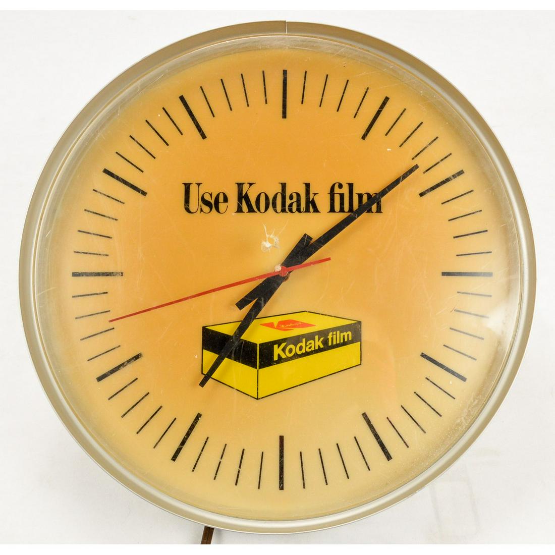 Kodak Film Lighted Advertising Clock: A lighted clock advertising Kodak film with the clock face marked "Use Kodak film" and a picture of a box of Kodak film. Clock has hour, minute, and second hands. Plastic covering has a small hole nea