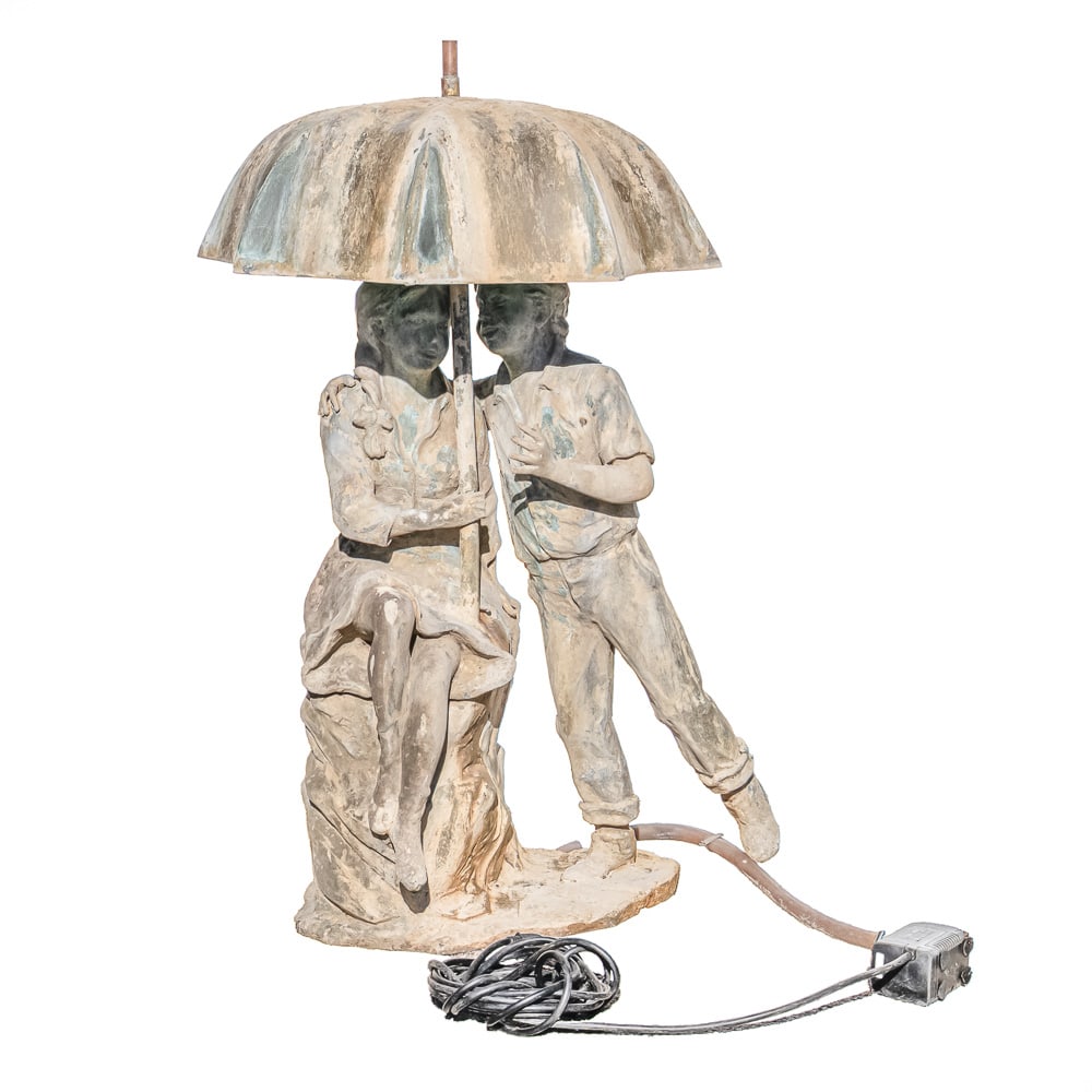 Boy and Girl Umbrella Fountain: A patinated metal garden fountain sculpture depicting a boy and girl standing together under a large umbrella. The figures are positioned on a naturalistic rockwork base, with the girl seated and the 