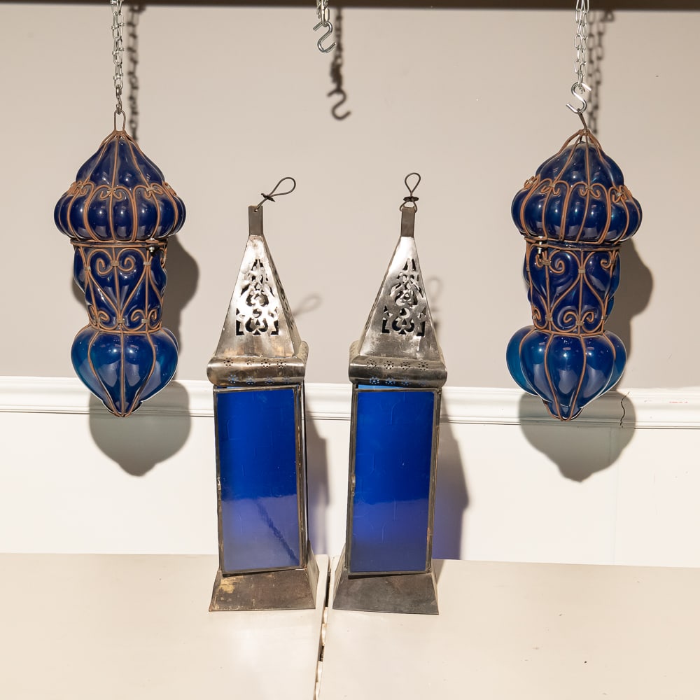 Blue Glass Moorish Style Lantern Set(4) w/4 Rods: This striking collection of four Moroccan-inspired lanterns captures the exotic flair of the late 20th-century revival of Moorish design. The set features two tall, obelisk-shaped lanterns with