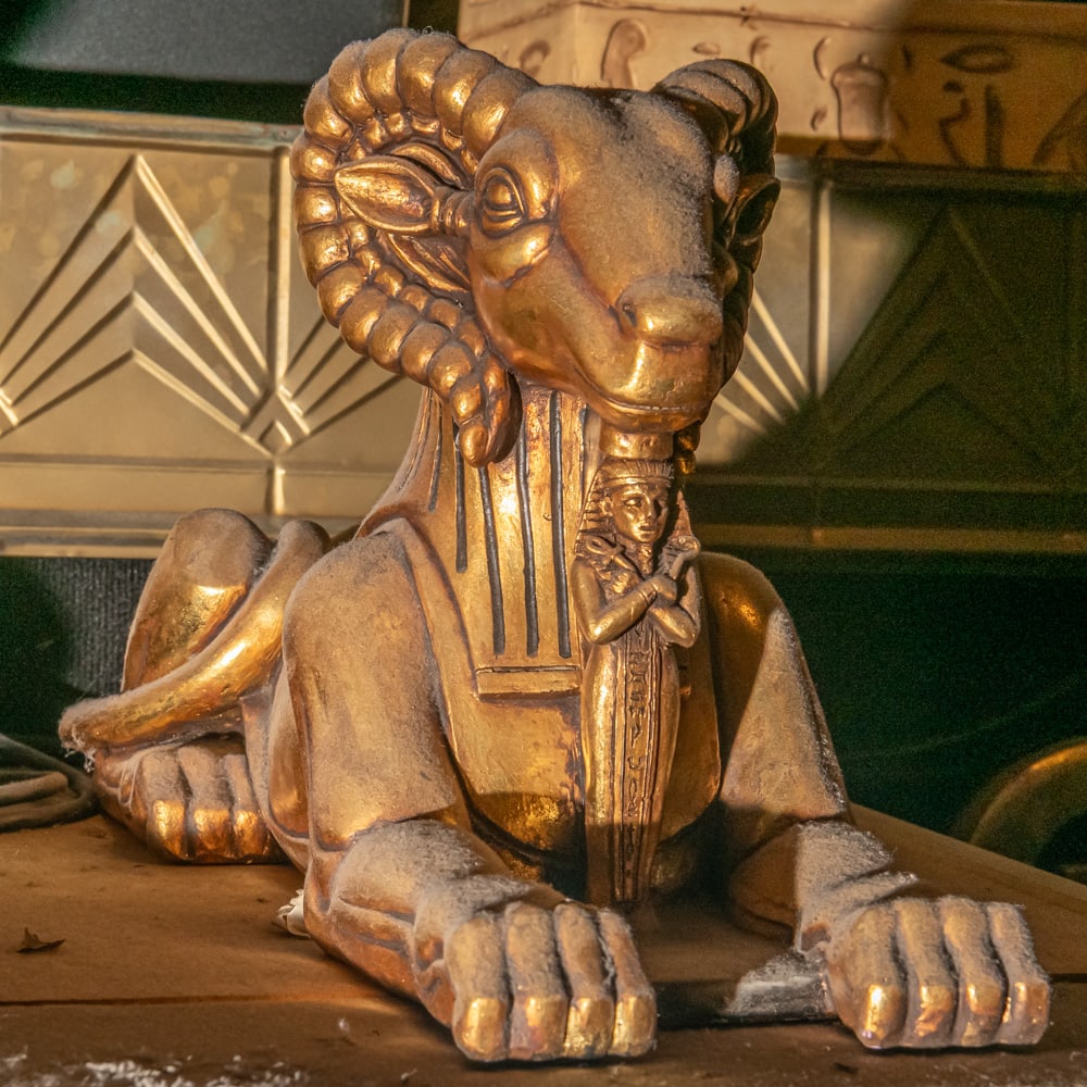 Sphinx-like Ram Carved Statue Right: Majestic recumbent ram statue featuring large eye catching carved horns. Ram lies in sphinx like divine protective pose with hind legs extended while the front ones are folded. This is the right side