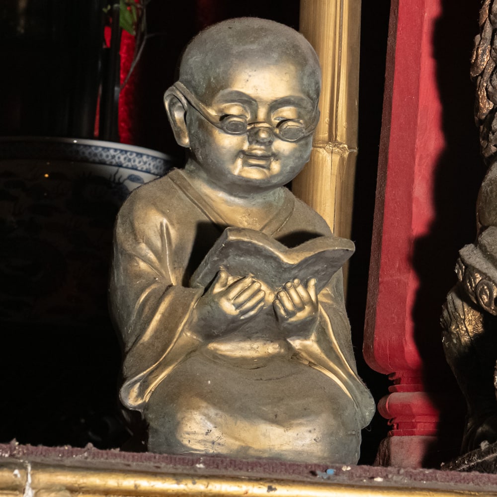 Scholar Monk with Spectacles Figurine: This charming figure of a young scholar-monk captures a whimsical moment of study, likely crafted from a cast resin or composite material designed to mimic the weight and patina of aged bronze. The pi