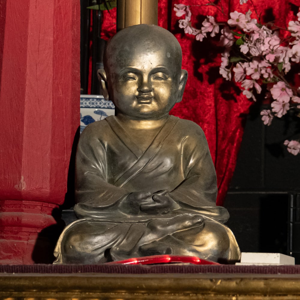 Bronze Patinated Seated Monk Statue: This serene, cast metal figure depicts a young Buddhist monk in a traditional seated meditation pose, hands resting in the Dhyana Mudra. The piece exhibits a rich, dark bronze-toned patina with subtle