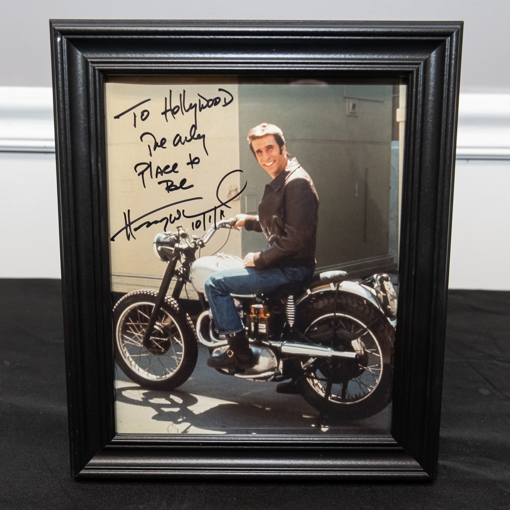 Framed Signed Henry Winkler "The Fonz" Photo 8x10: This classic piece of Americana features Henry Winkler as Arthur "The Fonz" Fonzarelli from the hit sitcom Happy Days, perched on his iconic Triumph motorcycle. The 8"x10" color photograph includes a