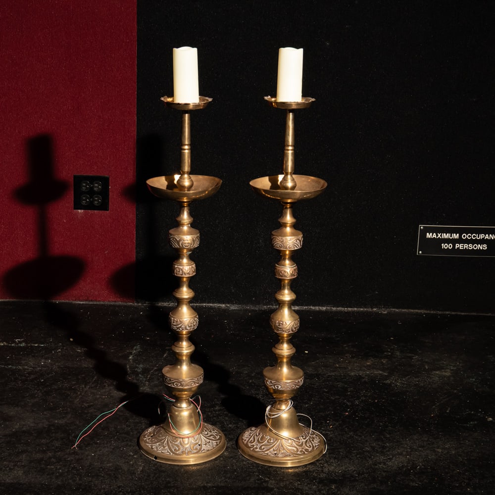 Pair Electrified Church Candle Stick Holders: This striking pair of tall, electrified candlesticks features a classic tiered baluster design with intricate repouss?-style floral and geometric patterns throughout the stems and flared bases. Likely