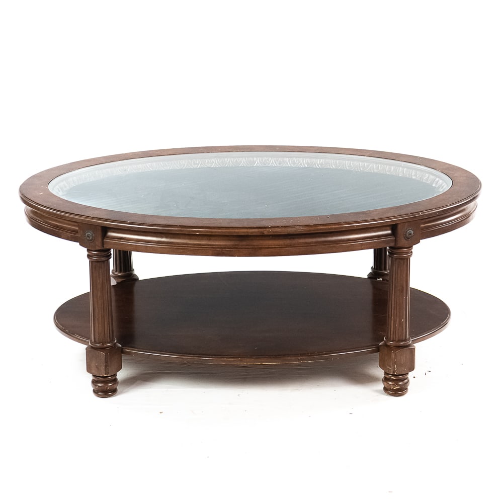 Oval Cocktail Table: Oval wood top with oval glass inset, shelf below, on legs. In good condition. Measures 48" x 32" x 19"Note: Pickup/ Transport required. Contacts available post auction.