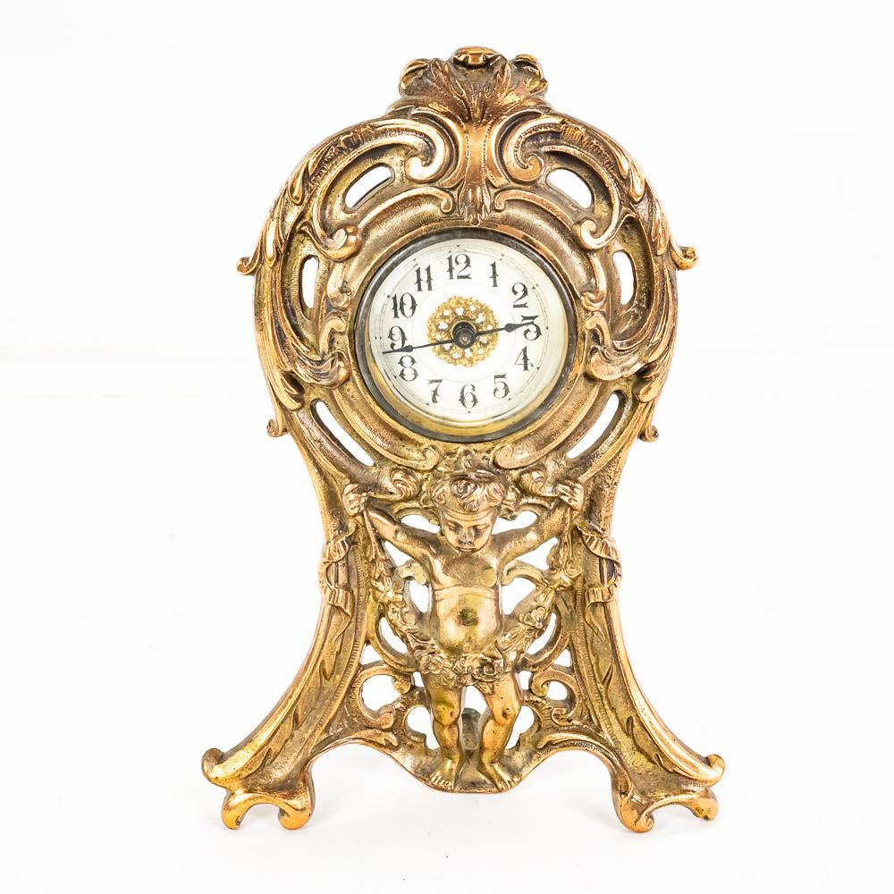Art Nouveau Cherub Gilt Mantel Clock: This is an evocative example of turn-of-the-century Art Nouveau design, featuring a central winged cherub or "putti" supporting a circular dial within a frame of flowing, organic scrollwork. The piece