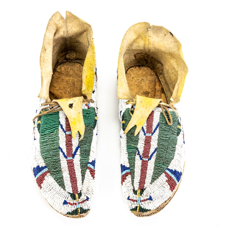 Antique Plains Fully Beaded Moccasins Pair: These exceptional Great Plains moccasins, likely Lakota (Sioux) or Cheyenne from the 1890–1910 period, feature masterful "lazy stitch" beadwork on brain-tanned deer hide. The central "feather" o