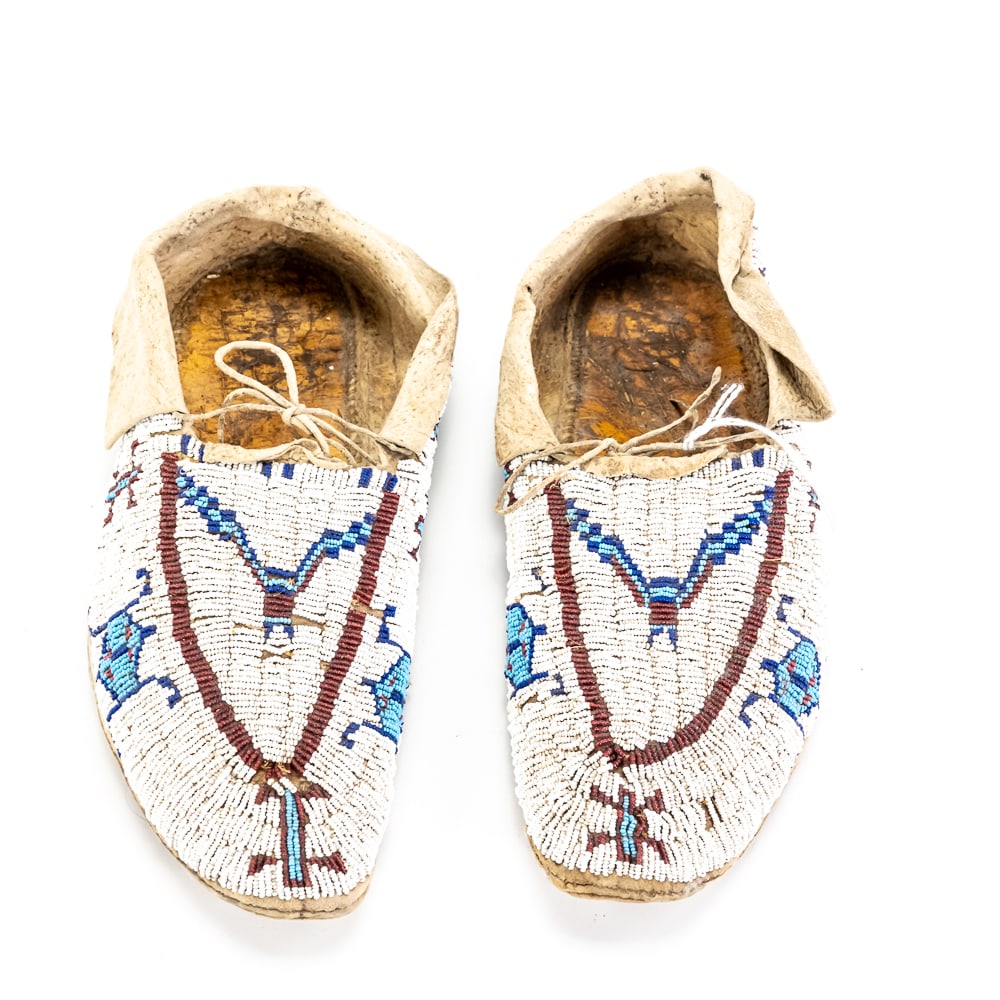 Native American Beaded Leather Moccasins Pair: These striking Native American moccasins are a testament to traditional Plains-style craftsmanship, likely dating to the early-to-mid 20th century. Hand-constructed from brain-tanned buckskin, they
