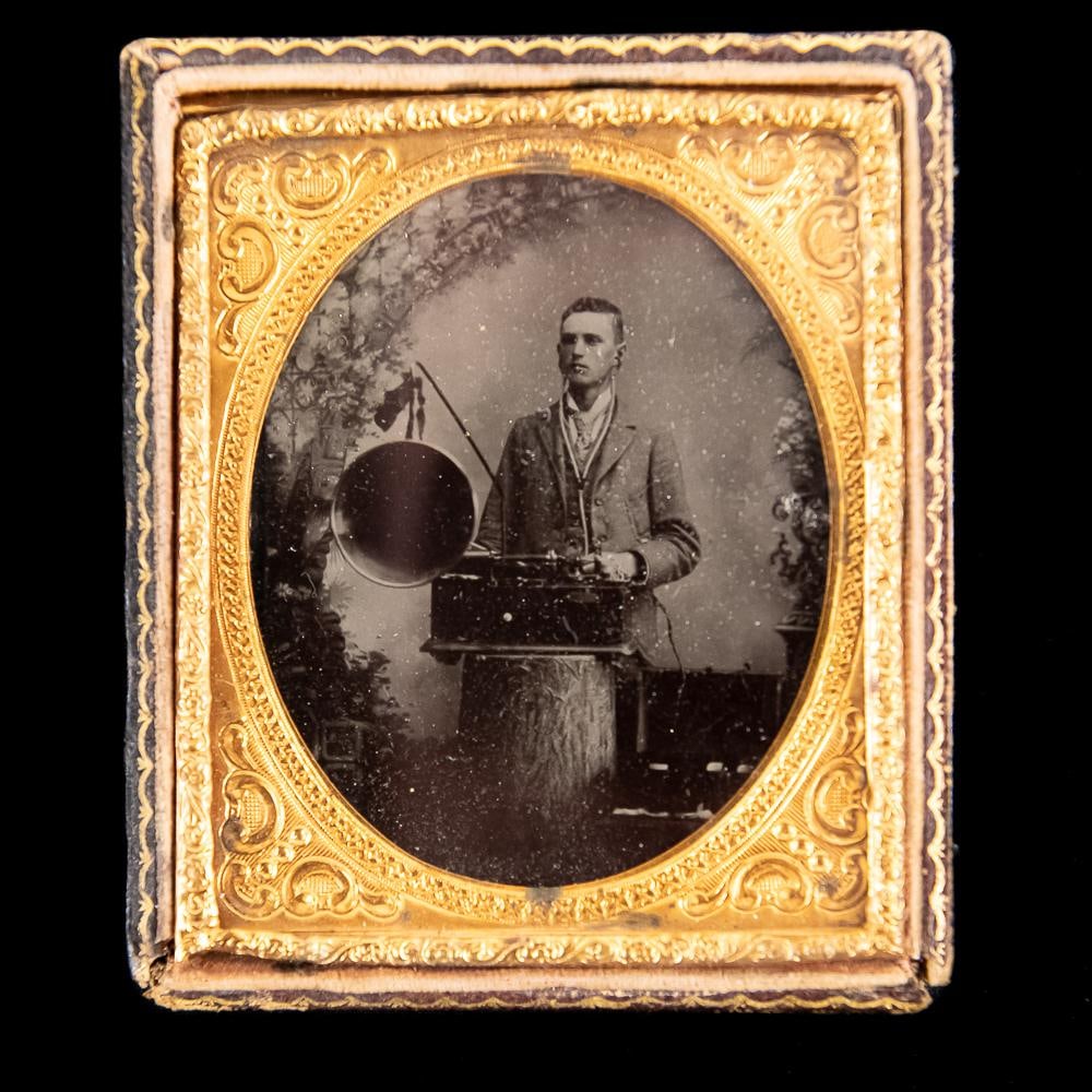 Tintype Of An Edison Class M Phonograph: 1/6 plate tintype image of a man standing behind an Edison Class M phonograph. The battery pack is visible in the lower right. In VG condition. From the Allen Koenigsberg collection