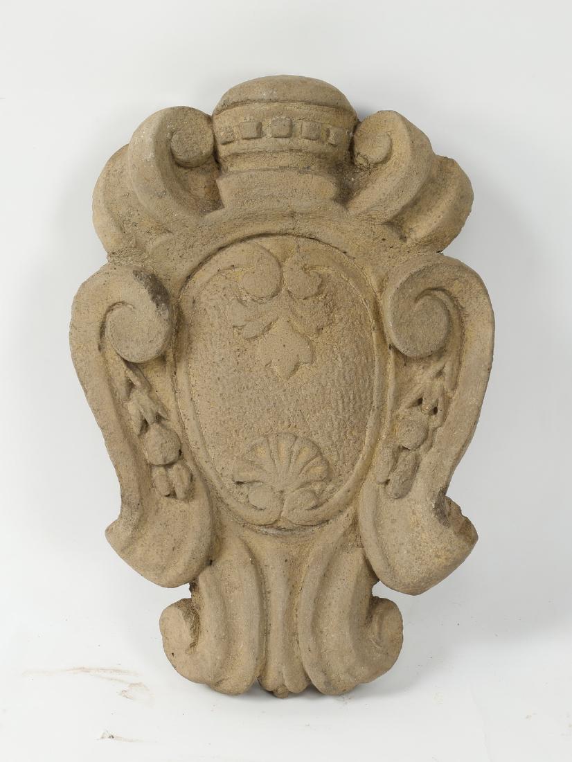 Continental heraldic crest in cast stone, 17"h: Cast stone Continental heraldic crest, having a geometric shaped crest flanked by scrollwork, surmounting a central shield decorated with a foliate palm and shell, finished with a scrollwork shaped bo