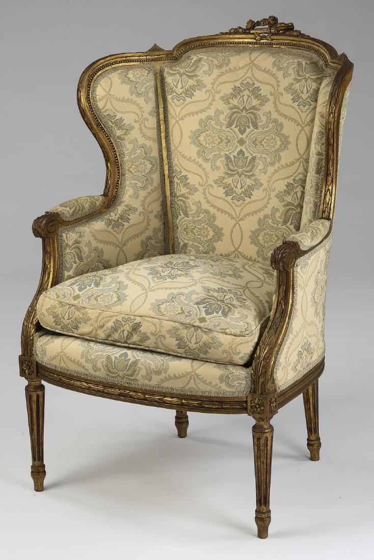 Carved giltwood bergere in the Louis XVI taste: Carved giltwood bergere in the Louis XVI taste, late 19th or early 20th century, having a carved floral torch crest above a jacquard upholstered back and wings flowing into partially upholstered arms,