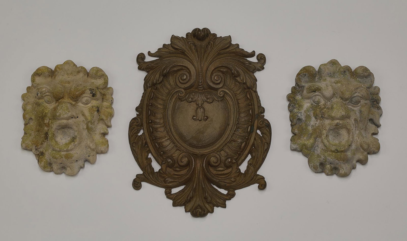 (3) Italian appliques,12"h and 7"h: Group of (3) Italian decorative appliques, including (2) cast stone grotesque masks, 7"h x 5.5"w x 2.25"d, and (1) bronze acanthus scrollwork medallion, 12"h x 8.75"w x 0.5"d.