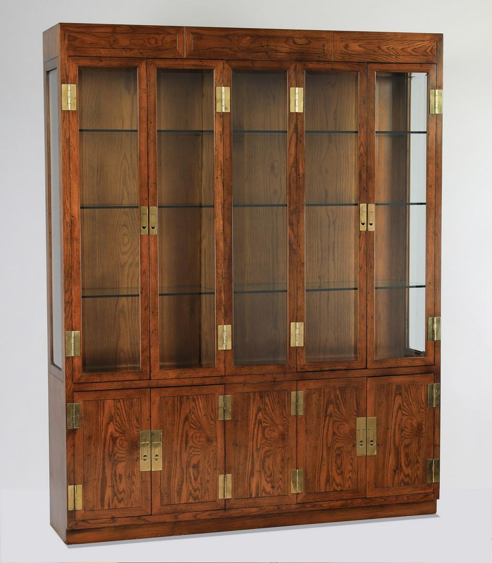 Henredon campaign style vitrine w/ beveled glass: Henredon campaign style oak and glass vitrine, having a pair of beveled glass doors on either side of a beveled glass panel, opening to a lighted interior with three glass shelves, flanked by beveled 