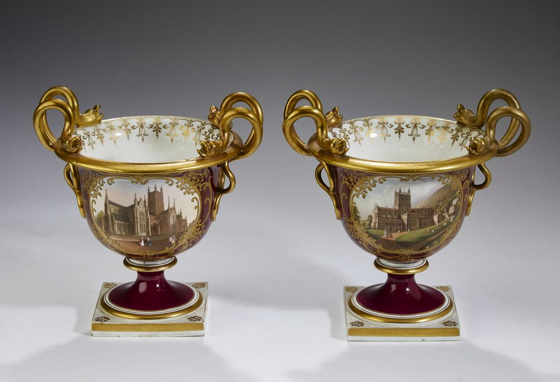 Pair of porcelain urns with snake handles, 8"h: Pair of porcelain urns, each having intertwining snake double handles and having a reserve depicting a Gothic church on a raspberry ground, rising on a socle base terminating in a square plinth, havin