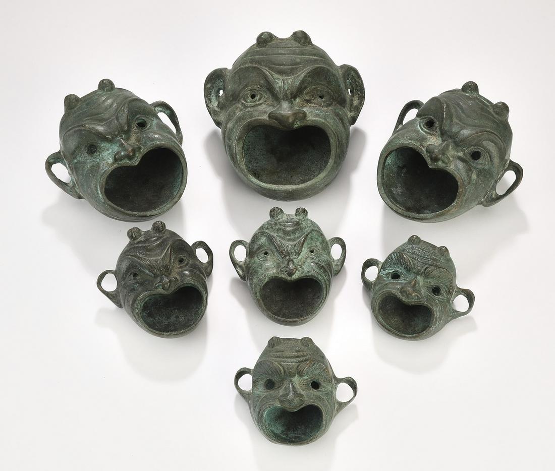(7) Ancient form, bronze figural hand held oil lamps: Group of (7) bronze hand-held oil lamps, depicting an expressive mask with stretched lobes and wide mouths, largest measuring 5"l x 4.5"w x 3"d, smallest measuring 2.5"l x 3"w x 1.5"d.