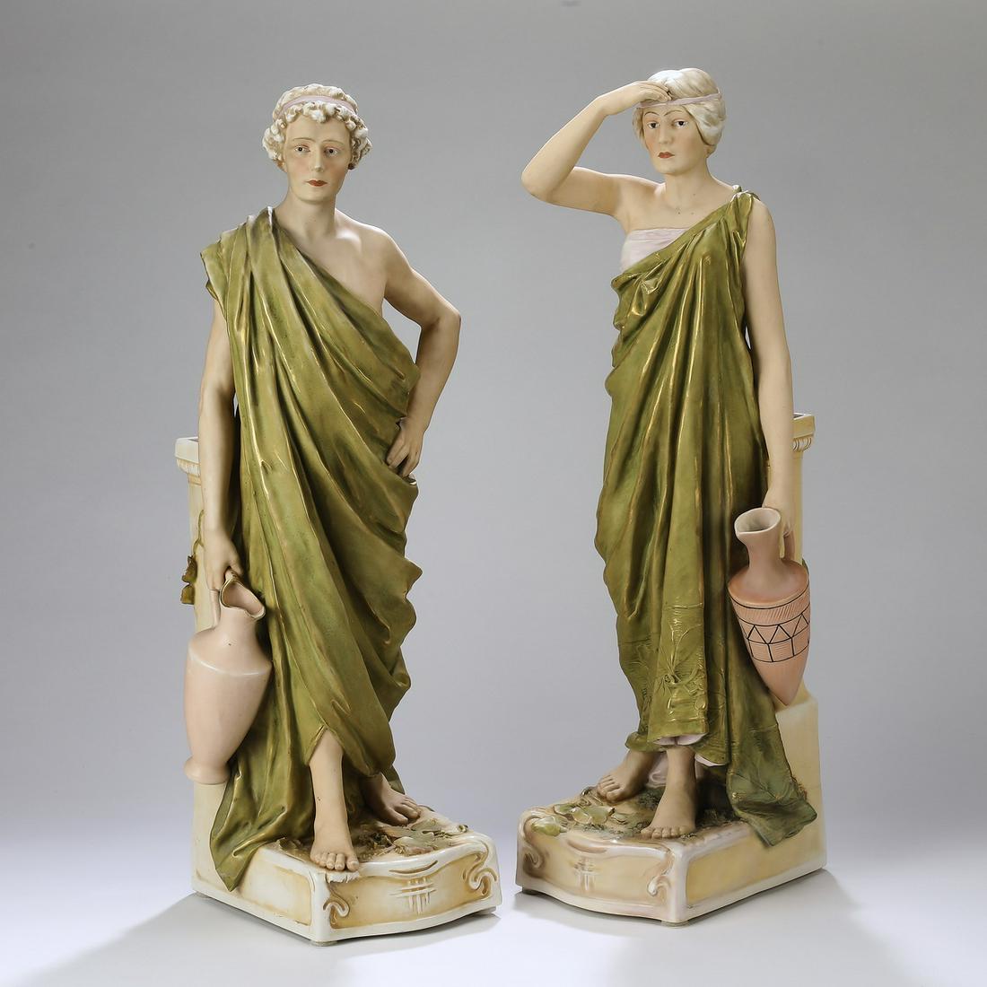 (2) Oversize Royal Dux figural vases, 25"h: Pair of oversize Royal Dux polychrome bisque figural vases, depicting male and female wine carriers, each dressed in a Classical toga, holding a wine jug, and leaning against a rectangular columnar va