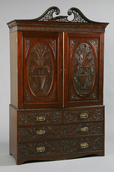 19th c. English mahogany linen press: Mid 19th century George III-style Cuban mahogany double door linen press with pierce carved broken arch pediment, over doors with relief carved oval panels featuring double handled urns filled with fo