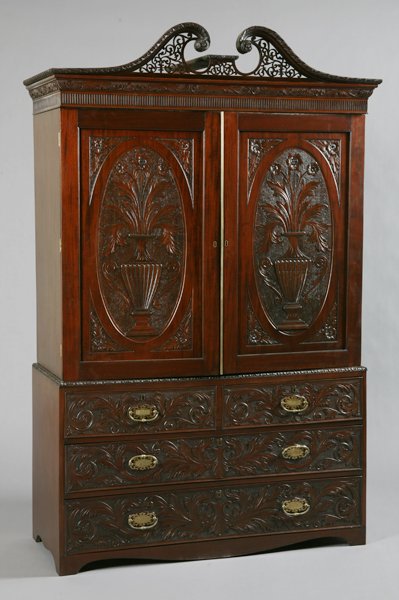 19th c. English mahogany linen press: Mid 19th century George III-style Cuban mahogany double door linen press with pierce carved broken arch pediment, over doors with relief carved oval panels featuring double handled urns filled with fo