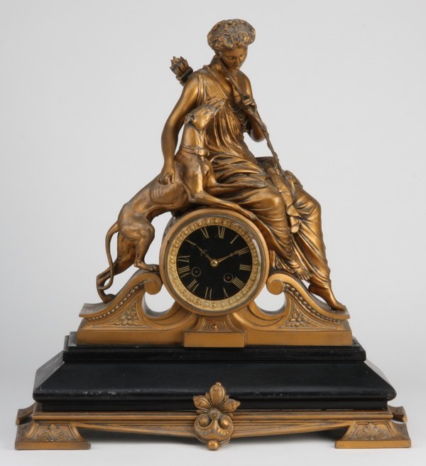 Early 20th c. Continental figural clock: Early 20th century Continental spelter and ebonized wood figural clock depicting Diana the Huntress with hound flanking the clock face, all surmounting a broken arched pediment raised on a shaped rect