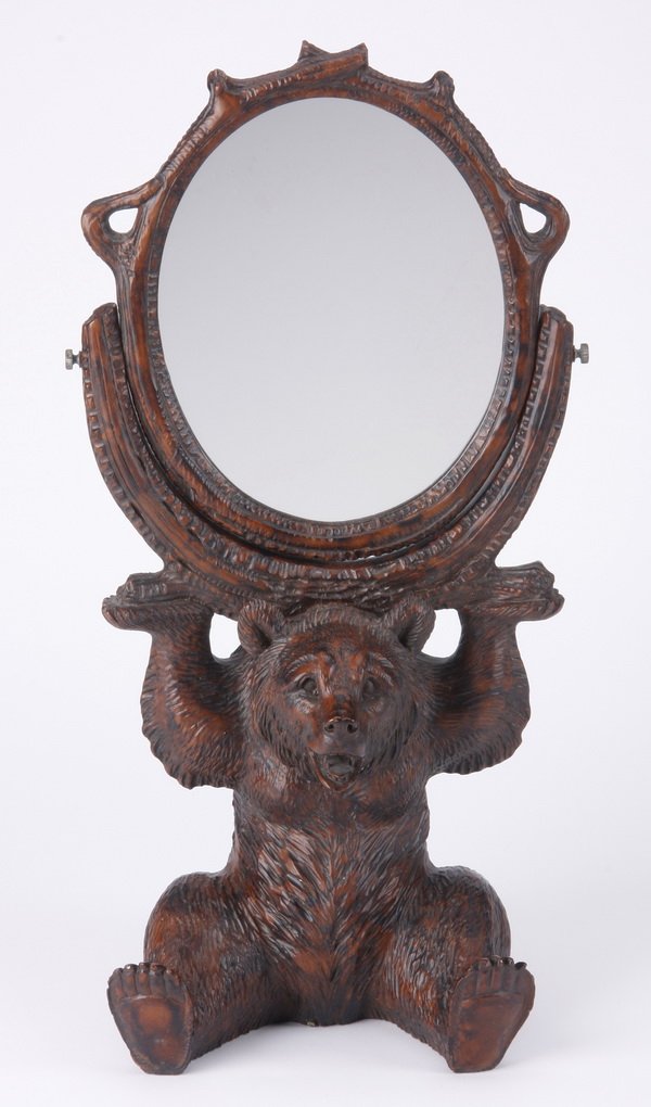 Black Forest inspired dressing mirror: Black Forest inspired dressing mirror of composite material, modeled as a seated bear holding aloft an oval mirror surrounded by a gnarled branch border, 20"h x 8.5"w x 7"d.