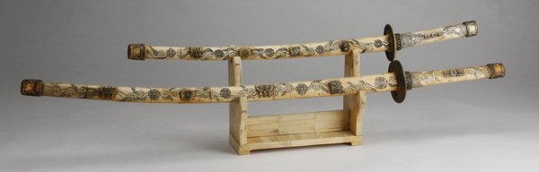 (2) Carved bone scabbards & sword handles: Two late 20th century carved bone decorative scabbards with sword handles and no blades, incised with floriate and foliate designs, with a corresponding stand, (1) 41"l, (1) 30.5"l, base 8.5"h x 10.25