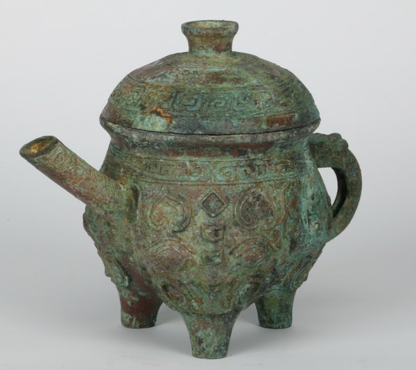 Chinese Warring States style bronze ewer: Chinese Warring States style he or ritual bronze ewer, the tripod vessel with a globular body cast with taotie designs, surmounted by a cover and raised on tapered feet, 10"h x 12"w x 8"d.