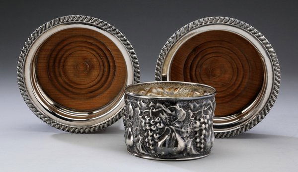 (3) Silver plate wine bottle coasters: Group of three silver plate wine bottle coasters: (2) with a gadroon border and wood liner, 6.25" diameter, and (1) Italian repousse with clusters of grapes, marked Delberti Italy, 3"h x 4.5" diameter