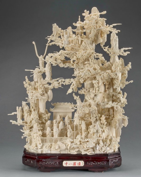 Chinese bone carving of 18 Louhan on sacred mountain: Chinese bone carving depicting the Eighteen Louhan on a sacred mountain with pagodas, parasol trees, flying cranes, and auspicious cloud forms, raised on a carved ebonized wood base with inscription, 