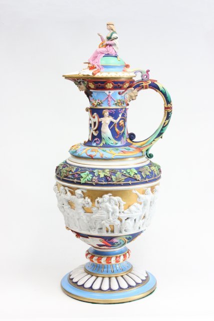 Magnificent 19th c. covered German porcelain ewer: Depicting "Drink Scene" around center. Adorned with faces. Includes seated figure on lid. Approx. 38" H. Base restored. Chip on interior lid. Bolt missing to hold lid to base.