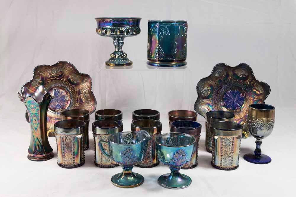 Group Lot of Blue Carnival Glass: 2 ruffled plates with dragon & flower design, approx. 8 1/2" diameter. Vase, approx. 6 1/2" H. 10 tumblers, sugar bowl, & creamer with grape and leaf design. Wine goblet, compote, biscuit jar. Total 1