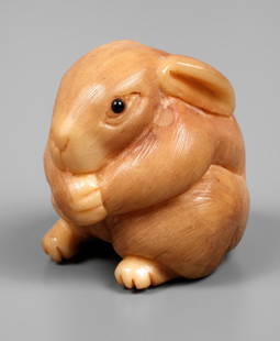 Netsuke: Japan, um 1920, Unterseite signiert, Bein? fein beschnitzt, eingesetzte Glasaugen, in Form eines sitzenden Häschens, H 3 cm.