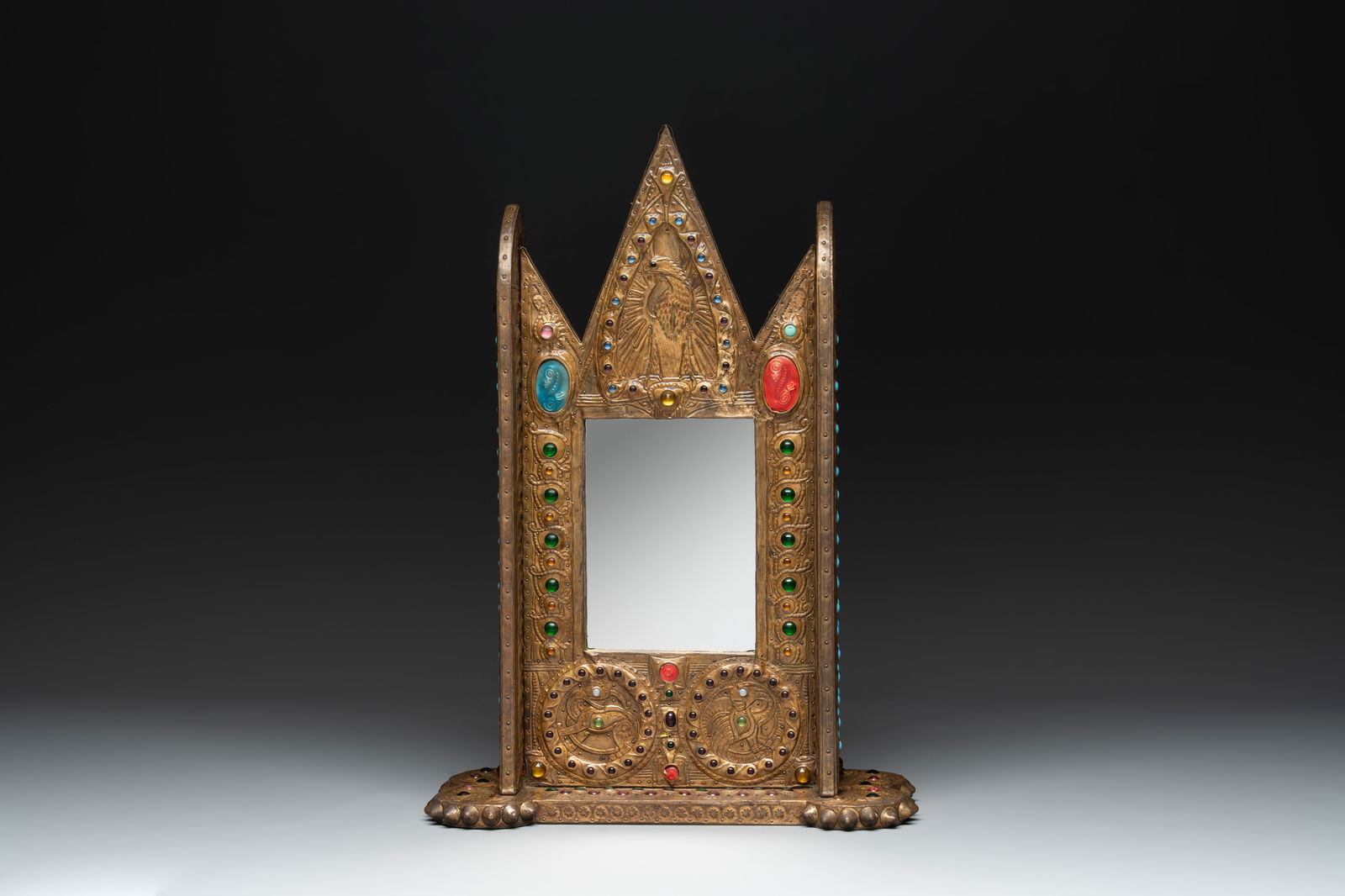 Alfred Daguet (Paris, 1875-1942): A Gothic Revival repoussé brass and copper-mounted mirror with: Alfred Daguet (Paris, 1875-1942): A Gothic Revival repoussé brass and copper-mounted mirror with glass cabochons, signed and dated 1907 Dim.: 32,5 x 15,5 x 46 cm Alfred Daguet was a French metalworke