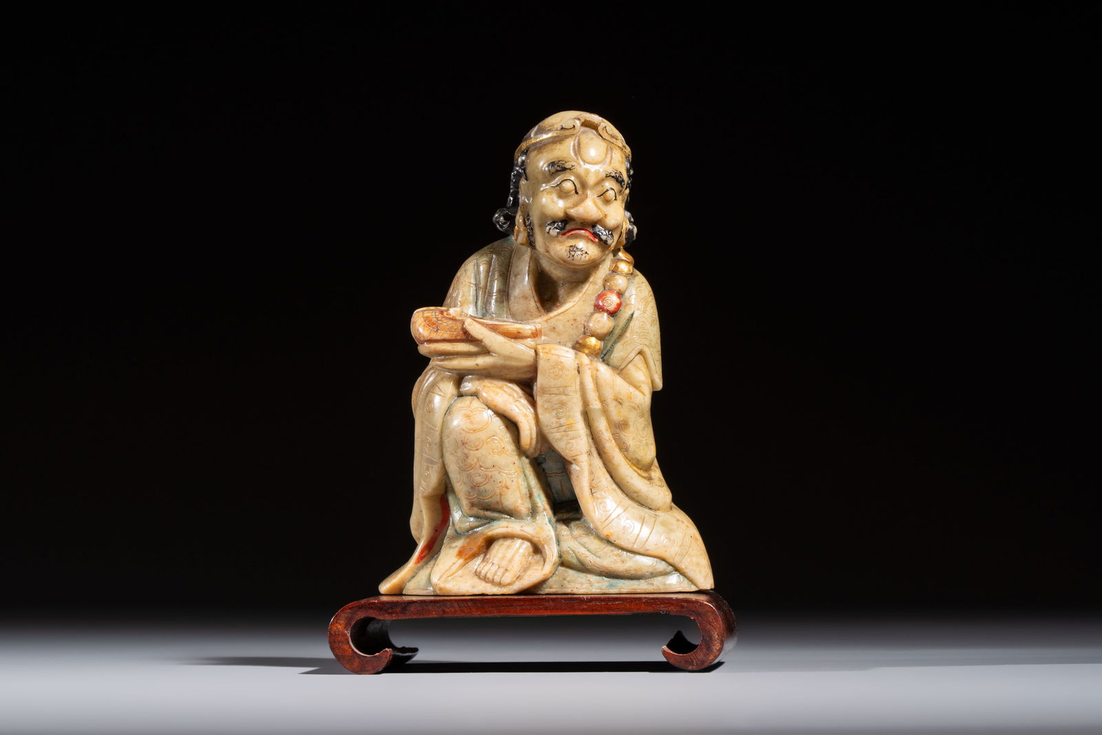 A Chinese gilt-polychromed soapstone Luohan on a wooden stand, 19th C.: A Chinese gilt-polychromed soapstone Luohan on a wooden stand, 19th C. H.: 13,2 cm (incl. stand) H.: 11,5 cm (excl. stand) Weight: 395 grams (excl. stand)