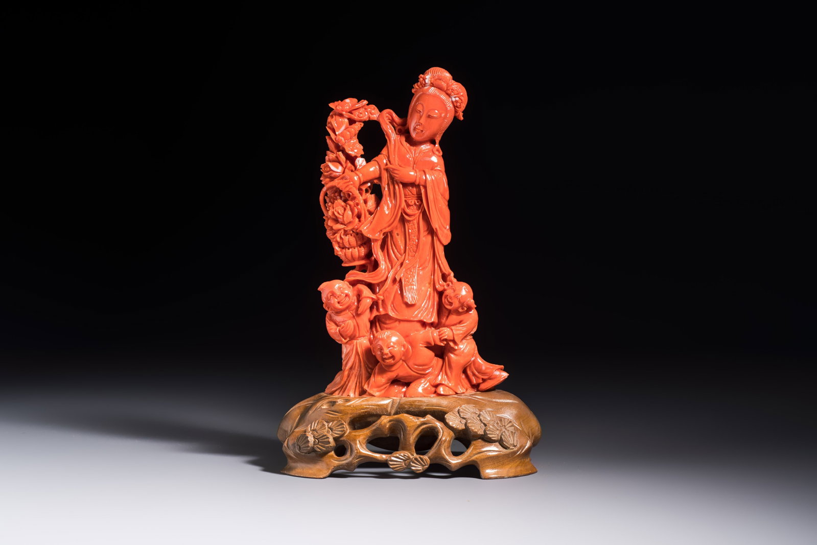 A Chinese carved red coral 'Goddess and Boys' group on a wooden stand, 19/20th C.: A Chinese carved red coral 'Goddess and Boys' group on a wooden stand, 19/20th C. H.: 19,2 cm (incl. stand) H.: 15,6 cm (excl. stand) Weight: 367 grams (excl. stand)