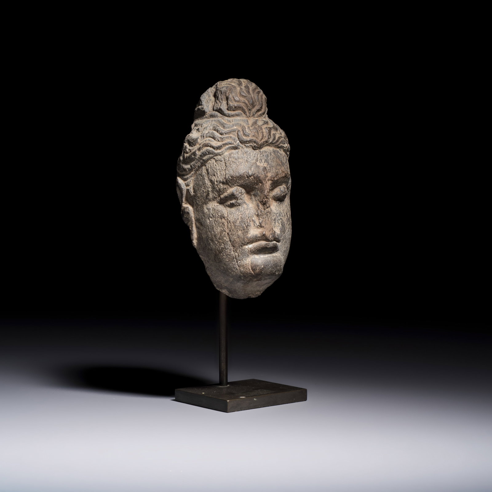 A Gandhara grey schist relief fragment of a Bodhisattva head, 3rd/4th C.: A Gandhara grey schist relief fragment of a Bodhisattva head, 3rd/4th C. H.: 22,6 cm | incl. stand H.: 15 cm | excl. stand